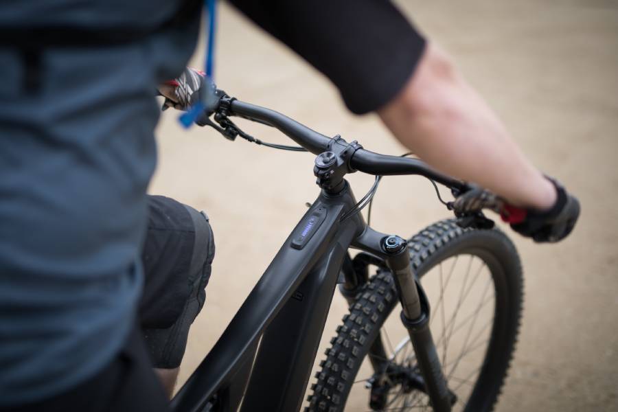 An over-the-shoulder, high-angle shot of a cyclist riding a matte black electric mountain bike on a dirt trail. The focus is on the wide handlebars, front suspension, and a digital LED display panel integrated directly into the top tube of the bike frame.