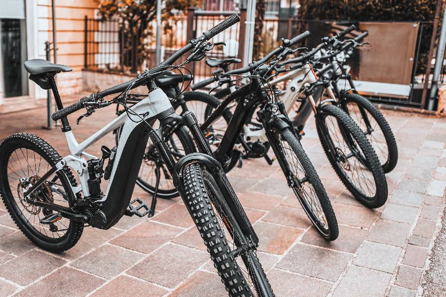 A fleet of several modern electric mountain bikes parked in a neat row on a stone-paved outdoor walkway. The closest bike in the foreground is a light grey and black full-suspension e-bike with thick, knobby tires.