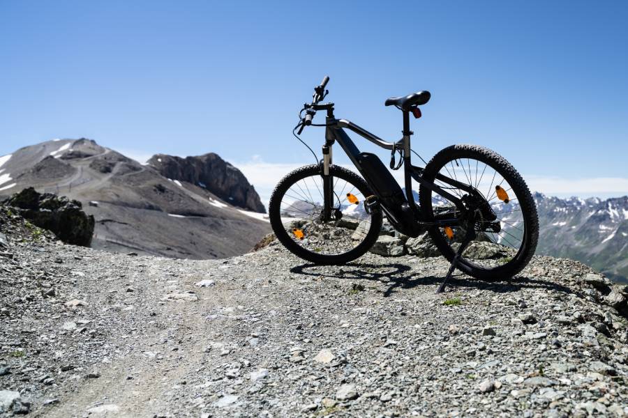 An electric mountain bike parked on a rocky, gravel trail, overlooking a vast panorama of rugged, snow-capped mountain peaks under a clear blue sky.