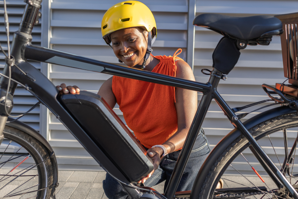 A woman in an orange top and bright yellow helmet smiling as she locks a removable black battery pack into the downtube of her electric bicycle outdoors.