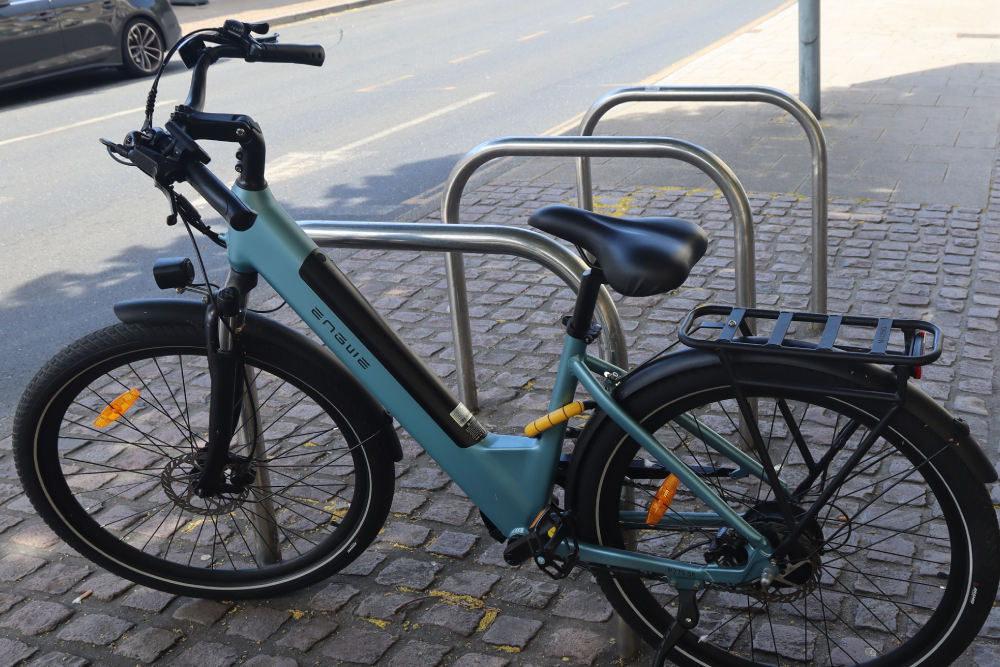 Light blue commuter e-bike equipped with essential accessories like a rear cargo rack, full fenders, and a headlight, parked securely on a street in Cambridge, Ontario.